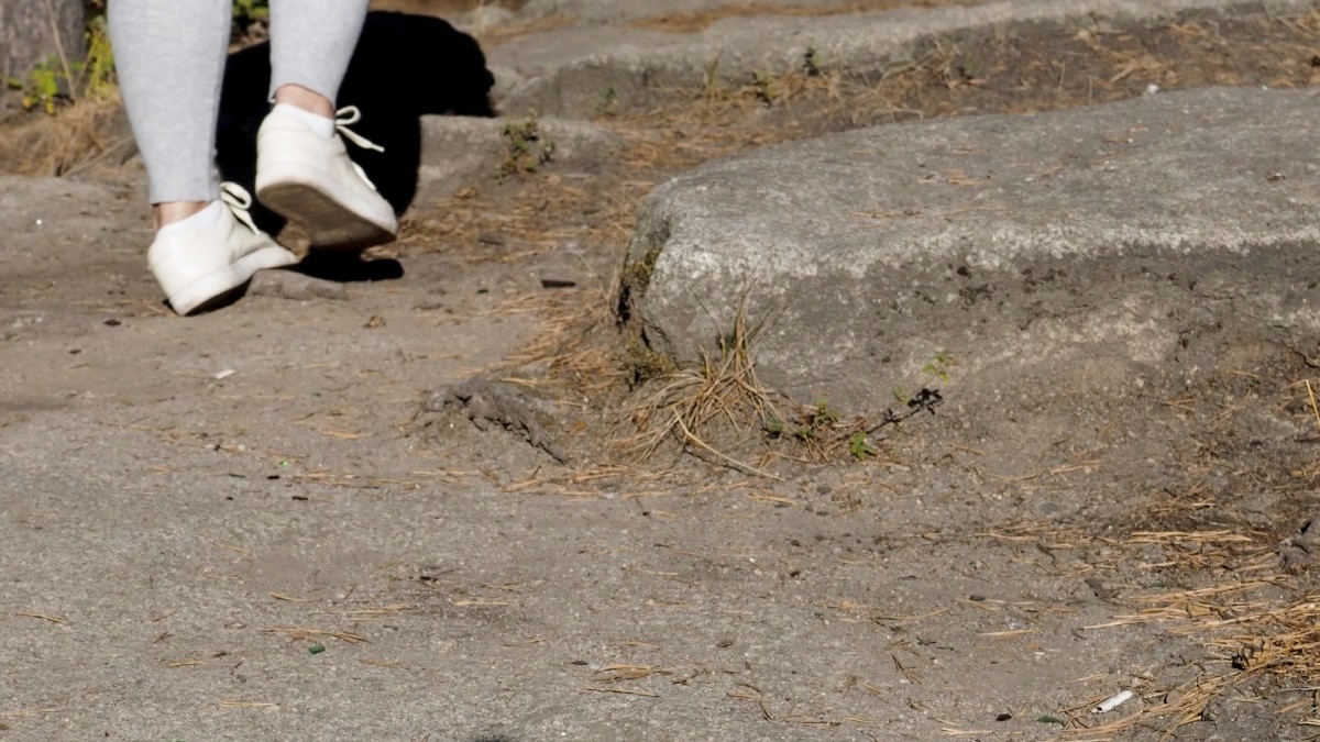 Walking Feet On The Asphalt. Motion. A Young Woman In White Sneakers And Jeans Walks Along A Sunny Road.